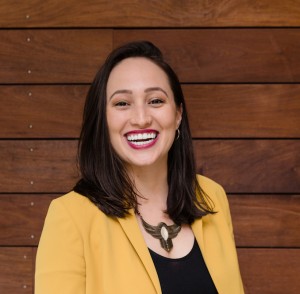 Alejandra smiles at the camera. She is wearing red lipstick and her hair falls just below her shoulders. She is wearing a yellow blazer with a black scoop neck shirt. In the background are horizontal wooden boards.